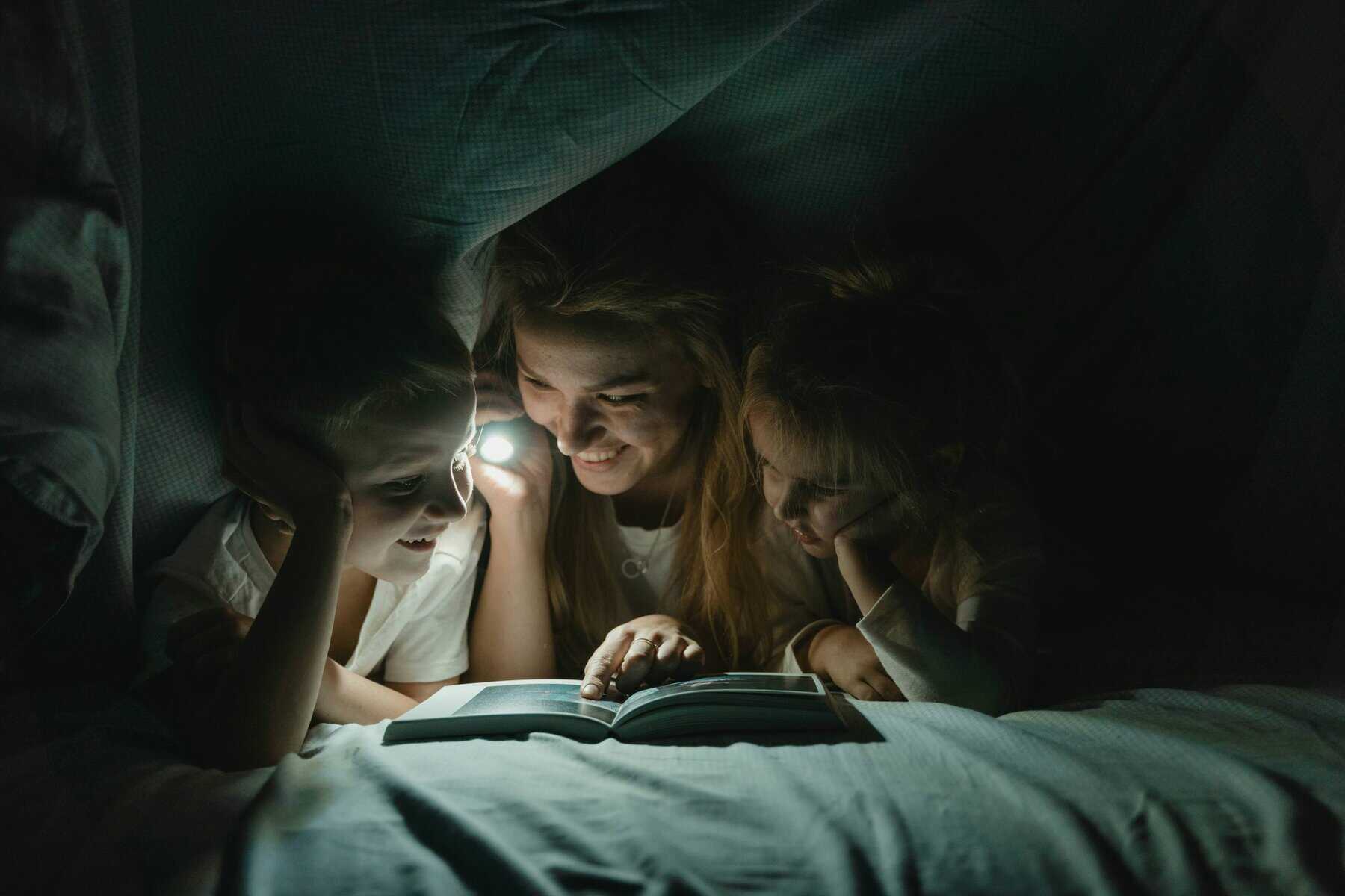 Mum and children reading a book by torchlight under blankets. Screen free time. Northants Families