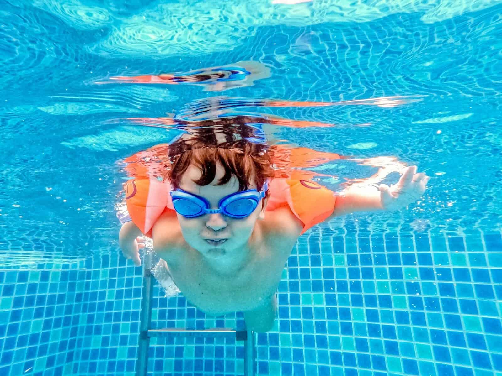 A child enjoying an underwater swim in a pool wearing goggles and arm floats.