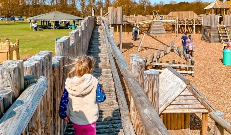 A child walks along a raised wooden walkway into a large timber playground on a sunny day.