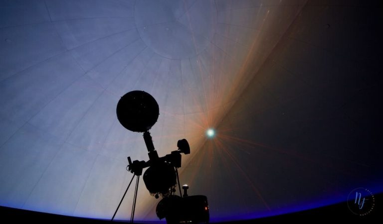 Silhouetted telescope-like instrument stands against a glowing sky with a sunburst and a thin blue horizon.