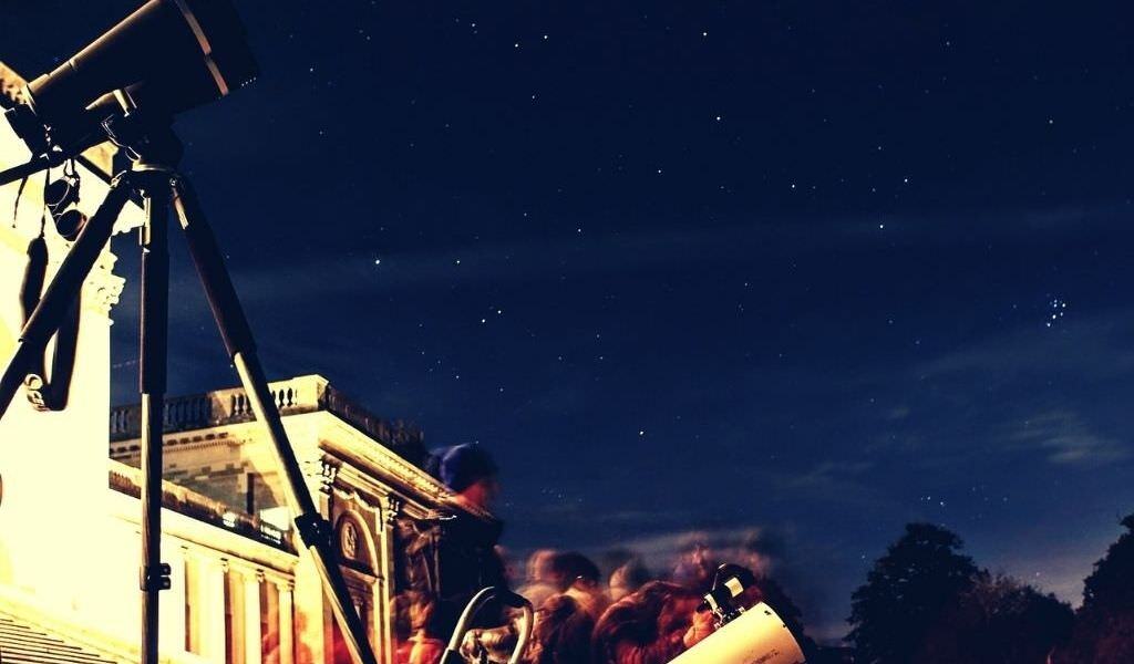 Public stargazing event with a telescope in the foreground and motion-blurred visitors beneath a starry sky.
