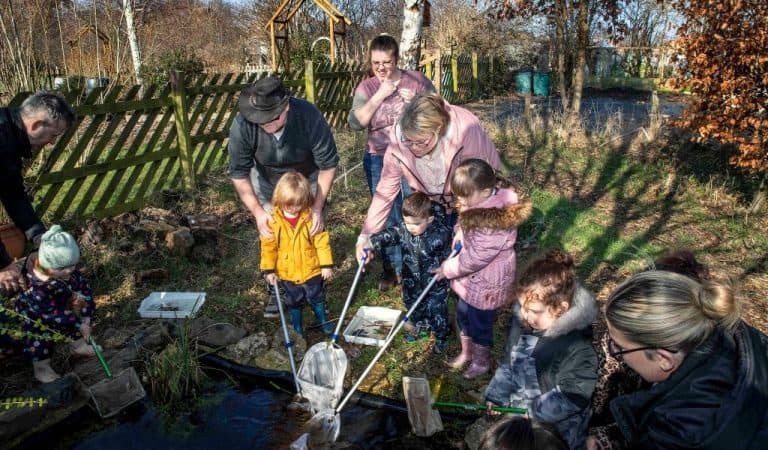 Adults and children gather around a small garden pond, using long-handled nets to scoop and inspect the water.