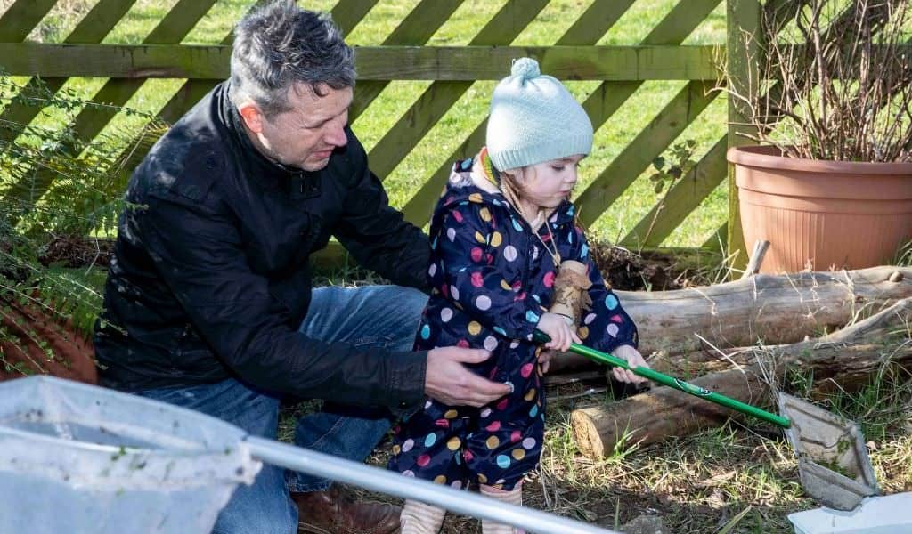 An adult kneels beside a child in a knit hat, guiding her as she holds a green-handled net near a garden pond.