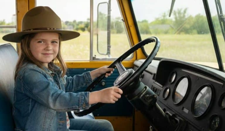 Little girl sitting at the steering wheel of an american school bus. She is wearing a ranger hat and appears to be in a field