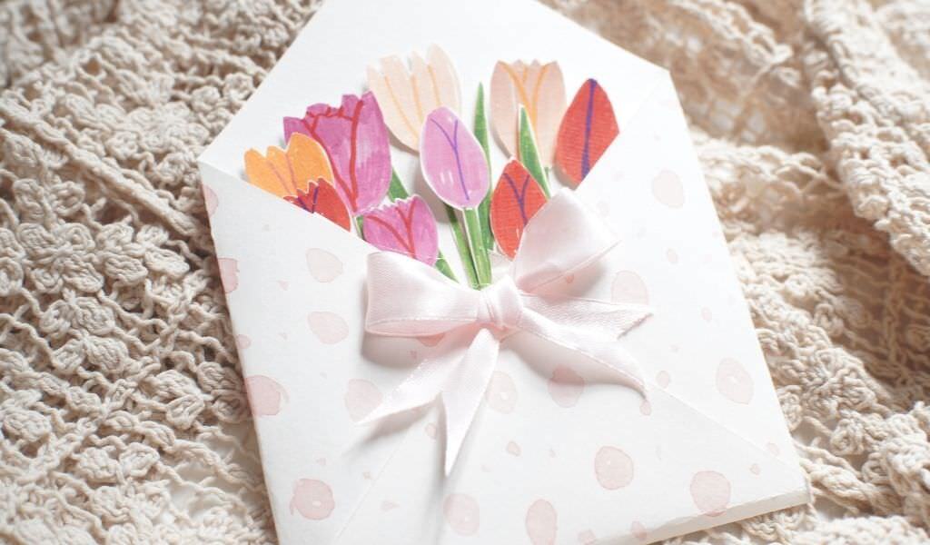 Handmade card with colorful paper tulips in a white envelope tied with a pink ribbon, resting on a knitted blanket.