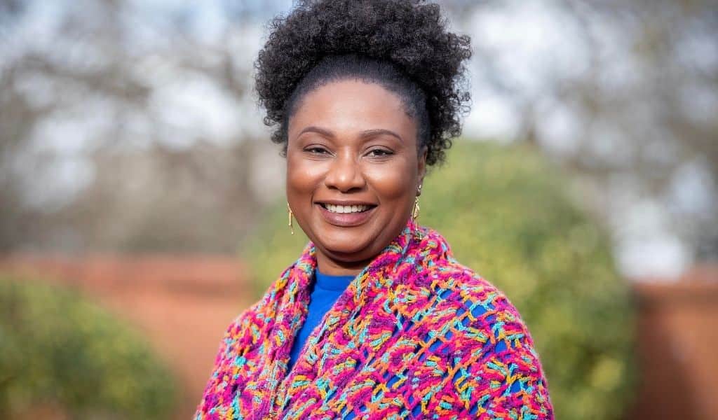Portrait of a smiling woman in a vibrant multicolored jacket, standing outside with soft-focus trees behind her and the “Bauhaus Education” branding across the image.