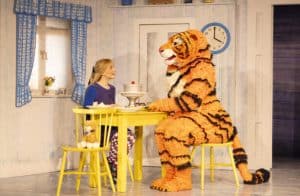 A scene from the theatre performance of The Tiger Who Came To Tea. A lady and the tiger are sitting at a yellow kitchen table talking over cake