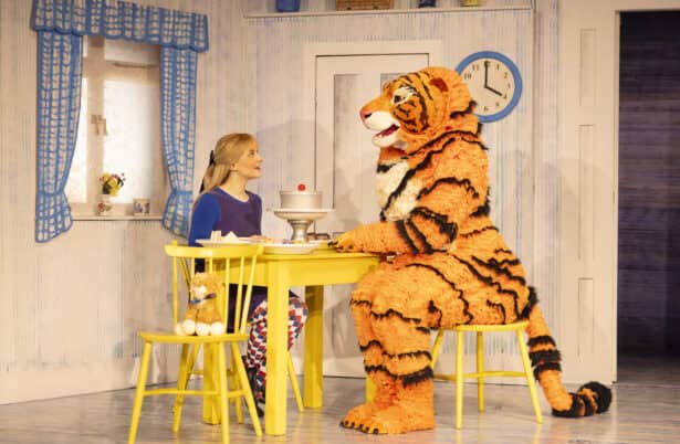 A scene from the theatre performance of The Tiger Who Came To Tea. A lady and the tiger are sitting at a yellow kitchen table talking over cake