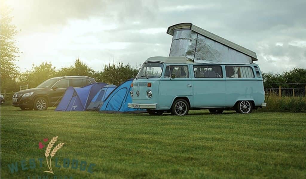 A camper van, two blue tents and a black car parked in a row in a field with the sun setting in the background