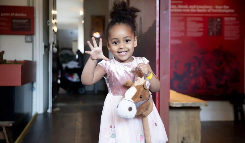 A little girl holding her open hand up to the camera standing ina room in Delapre Abbey. She is also holding a teddy in her left hand