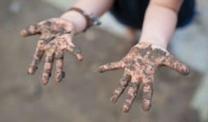 A young child holds out two hands covered in mud after playing outdoors, wearing dark shorts and a brown wristband.