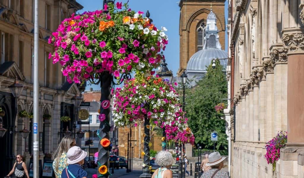 Hanging flower baskets on lamp posts in Northampton town centre with All Saints' Church tower in the background, part of the East Midlands in Bloom 2026 entry.