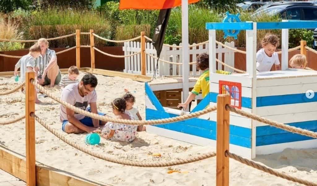 Dad and toddler playing in the sand at Rushden Lakes Beach, with children climbing on a wooden pirate-style play boat in the background on a sunny day.