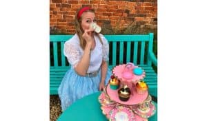 Performer dressed as Alice in a blue lace dress sits at an outdoor tea party table with a tiered cake stand of cupcakes and macarons, sipping from a floral teacup, ahead of Pitch Up Theatre's Alice in Wonderland at Lamport Hall