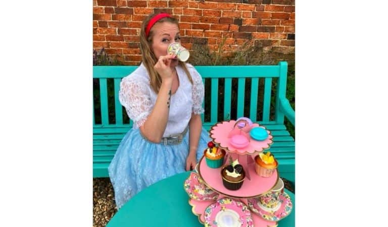 Performer dressed as Alice in a blue lace dress sits at an outdoor tea party table with a tiered cake stand of cupcakes and macarons, sipping from a floral teacup, ahead of Pitch Up Theatre's Alice in Wonderland at Lamport Hall