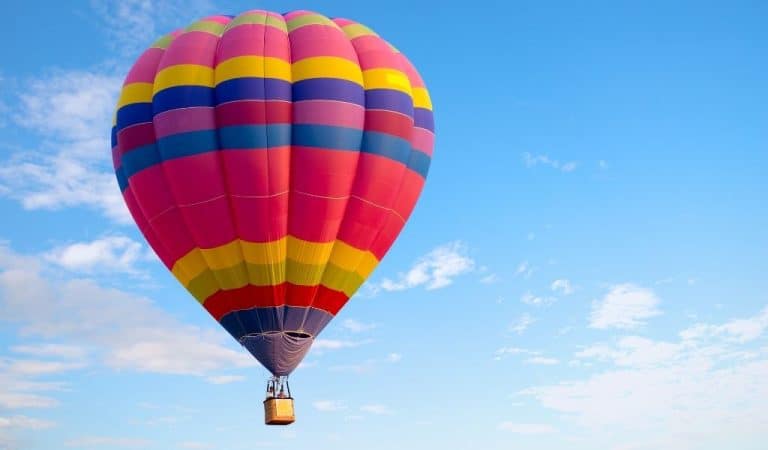 Colourful hot air balloon in a rainbow striped pattern flying against a bright blue sky with clouds, ahead of the Northampton Balloon Festival 2026