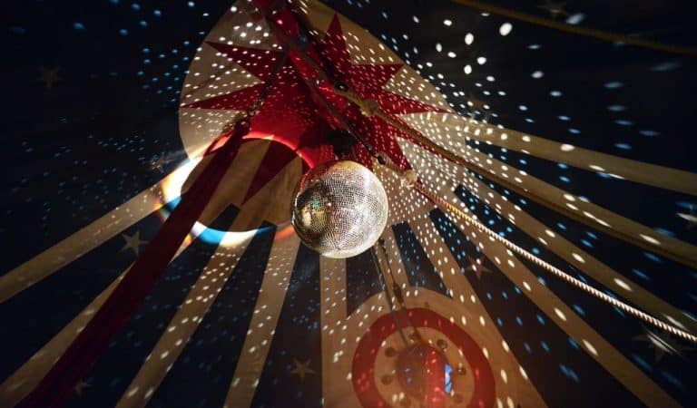 View up into a circus Big Top ceiling with red and cream star-patterned canvas, a sparkling disco ball catching the light, and rigging ropes stretching outward, lit with red and blue spotlights."