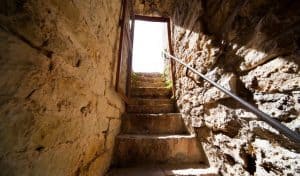 Stone staircase leading up through a narrow medieval passage to an open doorway, with a metal handrail alongside the worn steps