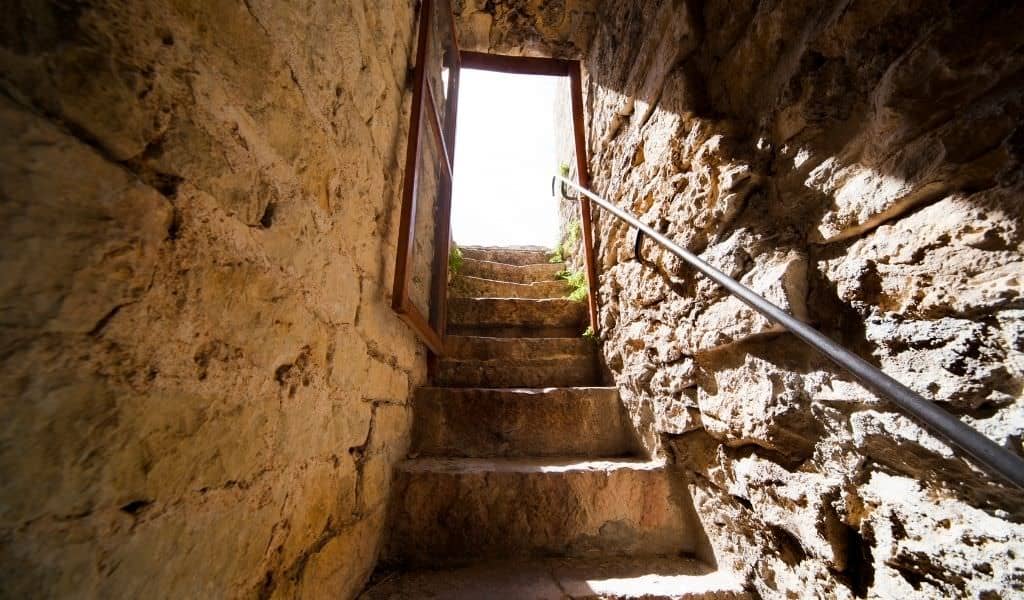 Stone staircase leading up through a narrow medieval passage to an open doorway, with a metal handrail alongside the worn steps