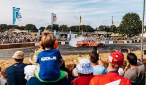 A young boy wearing ear defenders watches a stock car perform a smoky burnout on the track at CarFest, with crowds lining the barriers behind