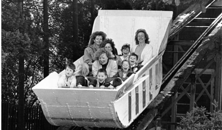 Black and white archive photo from the 1970s showing a group of women and children laughing and screaming with excitement as they ride down Wicksteed Park's Water Chute towards the splashdown