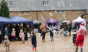 Children in winter coats and wellies watch a bubble maker in a red and white striped elf outfit send giant bubbles across the courtyard at Lamport Hall