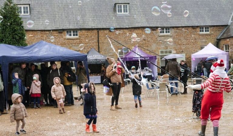 Children in winter coats and wellies watch a bubble maker in a red and white striped elf outfit send giant bubbles across the courtyard at Lamport Hall