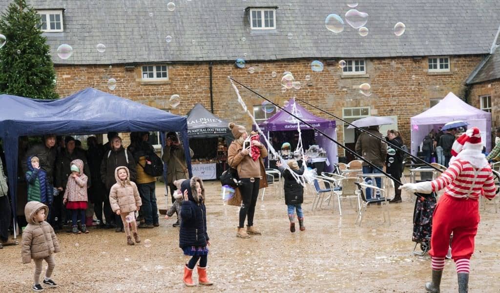 Children in winter coats and wellies watch a bubble maker in a red and white striped elf outfit send giant bubbles across the courtyard at Lamport Hall