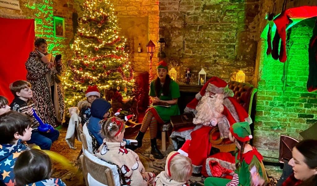 Father Christmas in a leather armchair beside a lit Christmas tree, with an elf reading to a group of children in a stone-walled room