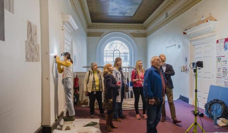 A small group of adults on a guided tour stand in a grand entrance hall at Stowe House, looking up at an ornately painted ceiling, while a conservator in protective gear works on a wall panel from a step stool nearby.
