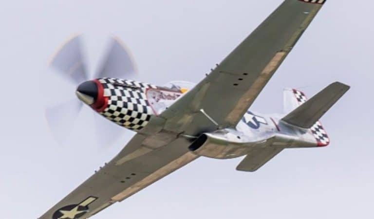 Vintage green P-51 Mustang fighter plane with black and white checkered nose banking sharply against a grey sky, propeller spinning.