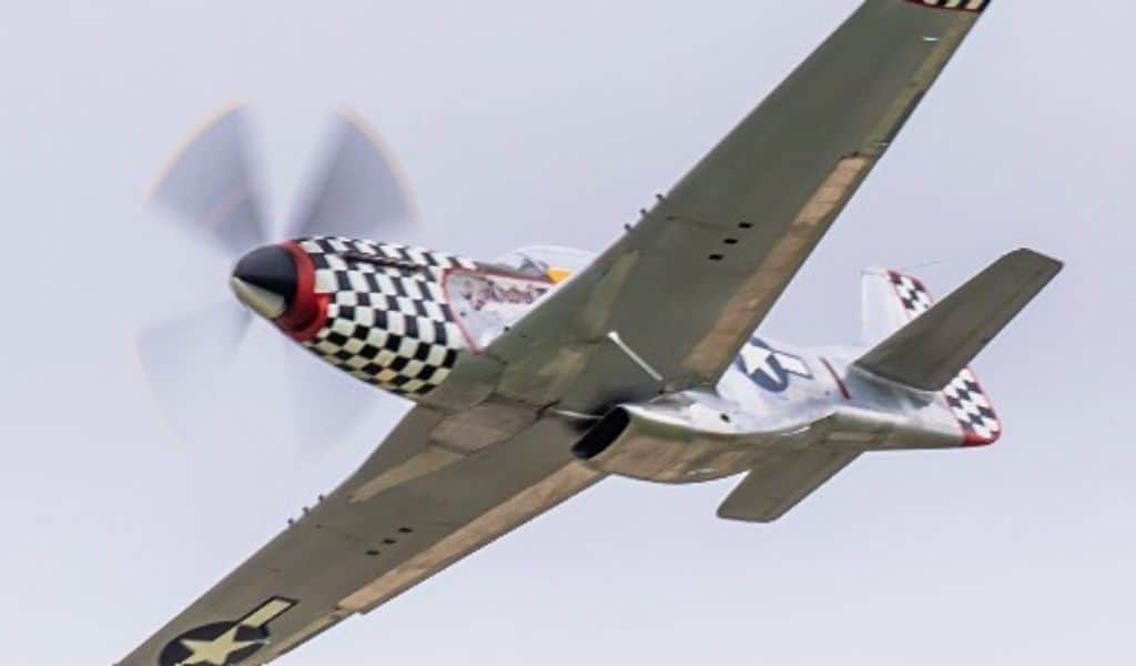 Vintage green P-51 Mustang fighter plane with black and white checkered nose banking sharply against a grey sky, propeller spinning.