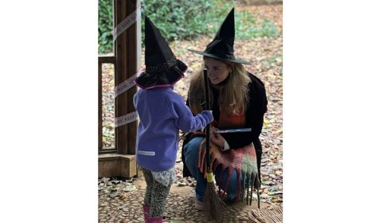 An adult in a black witch's hat crouching down to talk to a small child in a witch's hat and purple fleece, among autumn leaves in a woodland setting