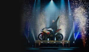 Illusionist performing a stage illusion on a motorbike surrounded by fireworks and stage lighting at an Illusion Impossible show
