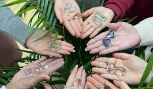 Several pairs of hands holding handmade resin jewellery pieces, including earrings and pendants set with dried flowers and glitter, next to a green potted plant