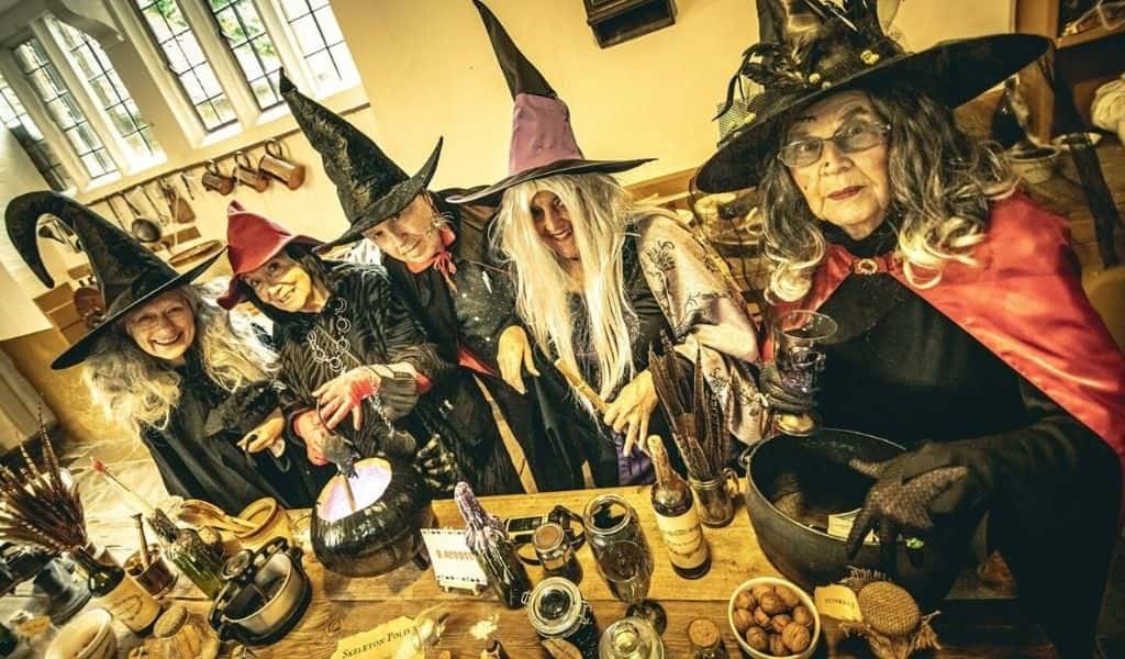 Five women dressed as witches in pointed hats gathered around a table with a cauldron and potion ingredients at Rockingham Castle's Halloween Fun event
