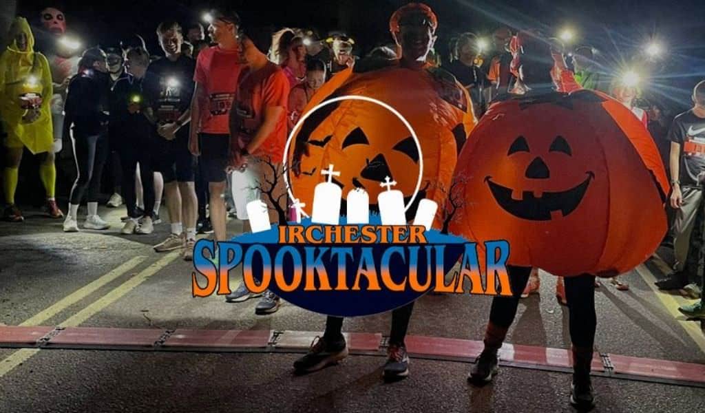 Runners in Halloween fancy dress, including inflatable pumpkin costumes, gather at the start line of the Irchester Spooktacular night run, with head torches lit in the dark and the event logo overlaid.