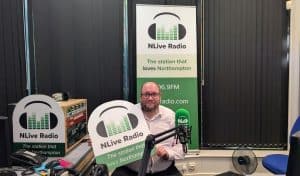 Man in glasses and a light pink shirt sitting at the NLive Radio studio desk holding an NLive Radio sign, with a green microphone and a banner reading "The station that loves Northampton" behind him