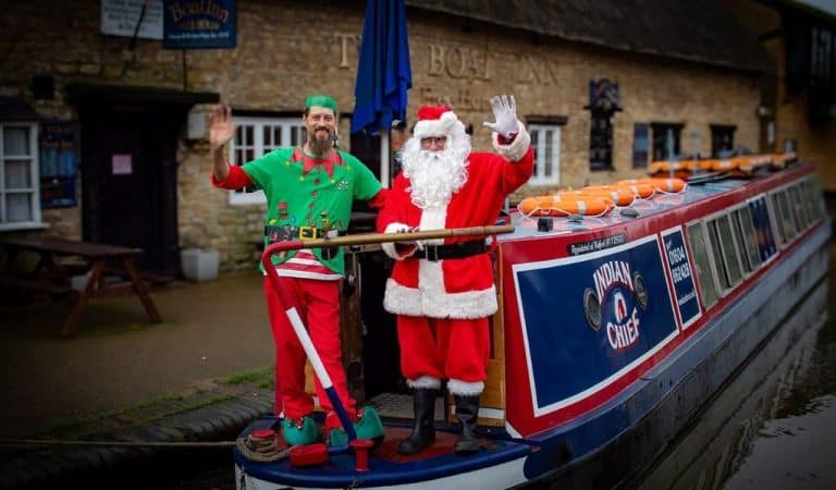 Santa and an elf in a green costume waving from the back of the narrowboat Indian Chief, moored outside The Boat Inn at Stoke Bruerne