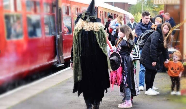 Families in Halloween fancy dress on the platform beside a red train carriage, with a witch walking past a little girl in a witch costume and a toddler in a pumpkin jumper