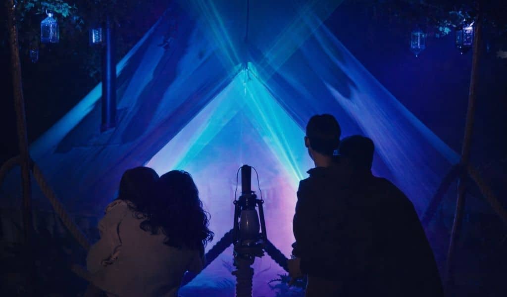 Families silhouetted in front of a tent lit with beams of blue and turquoise light, with a lantern in the centre, at the Forest of Secrets trail at Kelmarsh Hall